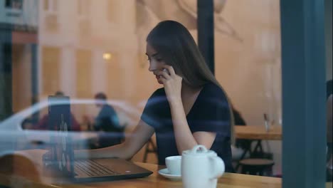 View-from-the-street-of-and-attractive-young-woman-talking-on-the-phone-in-a-cafe-and-looking-at-the-screen-of-her-laptop-during