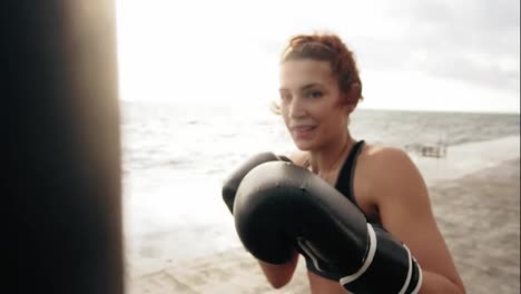Close-Up-view-of-a-strong-athletic-female-boxer-in-gloves-exercising-with-a-bag-against-the-son-by-the-sea.-Female-boxer-training