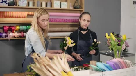 Attractive-blonde-florist-in-apron-standing-with-her-coworker-at-counter-in-floral-shot-while-arranging-bunch-of-flowers