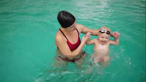 Young-mother-teaching-her-little-boy-how-to-swim-in-the-swimming-pool.-Baby-kicking-with-his-feet-in-the-water-to-start-swimming