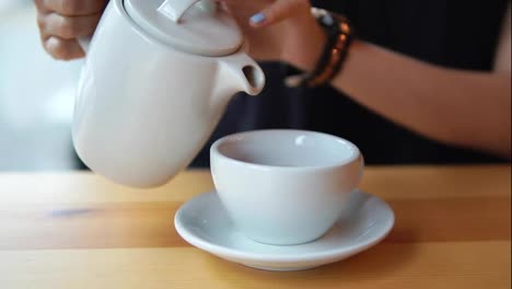 Close-up-view-of-female-hands-pouring-hot-tea-from-the-teapot-in-the-white-porcelain-cup-on-the-wooden-table-in-cafe