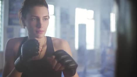 Close-Up-view-of-caucasian-female-boxer-hitting-the-boxing-bag-with-her-hands-in-gloves-in-the-gym-with-smoke.-Tough-power
