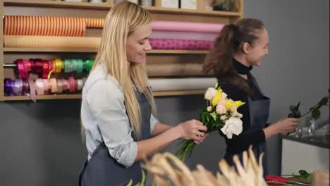 Smiling-blonde-florist-in-apron-standing-with-her-coworker-at-counter-in-floral-shot-while-arranging-bunch-of-flowers