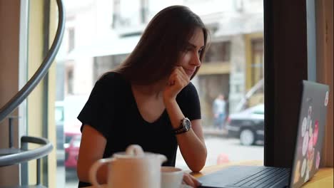 Young-woman-sitting-in-the-coffee-shot-by-the-window-having-a-coffee-break,-she-is-using-her-laptop,-working-and-surfing-in-the