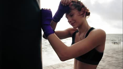 Close-Up-view-of-a-young-woman-having-a-break-after-hard-training-by-the-boxing-bag-against-the-son.-Her-hands-are-wrapped-in