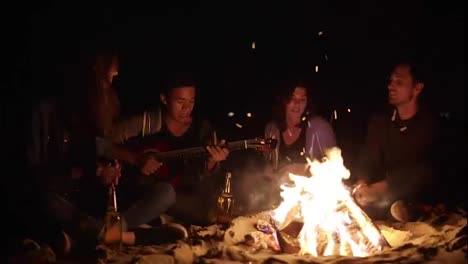 Multiracial-group-of-young-boys-and-girls-sitting-by-the-bonfire-late-at-night-and-singing-songs-and-playing-guitar.-Cheerful