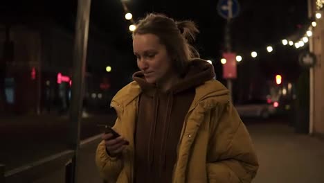 Short-haired-girl-walks-through-the-evening-city-smiling,-stops-and-texting-in-the-smartphone.-Handheld-footage