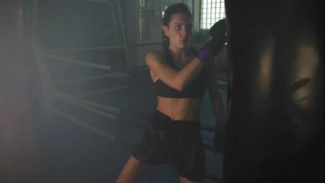Slow-Motion-shot-of-a-leg-hit.-Strong-athletic-female-kickboxer-exercising-with-a-boxing-bag-in-dark-gym-with-smoke-and-kicking-a