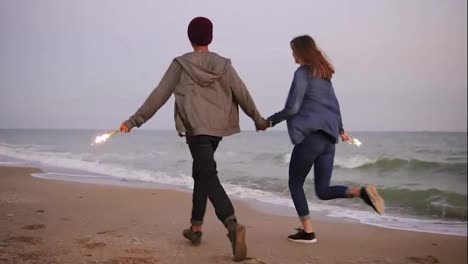 Back-view-of-young-multi-ethnic-couple-running-by-the-sea-and-holding-burning-sparkling-candles-during-sunset.-Slow-Motion-shot