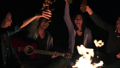 Group-of-young-people-near-campfire-drinking-beer-and-clicking-bottles-with-cheers-late-at-night.-Slow-Motion-shot