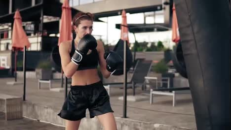 Attractive-athletic-female-boxer-in-gloves-kicking-a-punching-bag.-Workout-outside.-Female-boxer-training.-Self-defence-concept