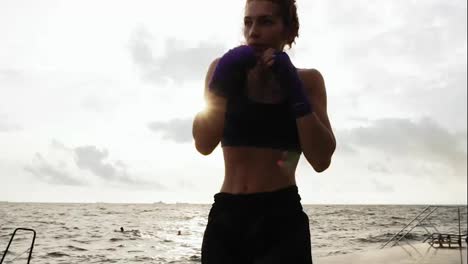 Close-Up-view-of-a-young-woman-shadowboxing-with-her-hands-wrapped-in-purple-boxing-tapes-by-the-sea-against-the-son.-Beautiful