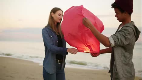 Young-multi-ethnic-couple-holding-red-paper-lantern-before-launching.-Romantic-date-on-the-beach.-Attractive-woman-together-with