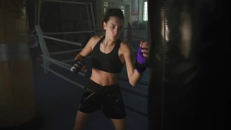 Athletic-female-boxer-in-gloves-hitting-boxing-bag-with-her-fist-while-training-in-a-dark-fitness-studio-with-smoke.-Slow-Motion
