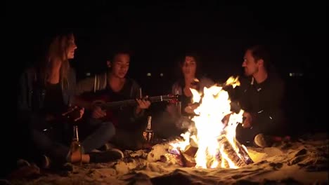 Cheerful-african-american-playing-guitar-for-his-friends-by-the-fire.-Multiracial-group-of-young-boys-and-girls-sitting-by-the