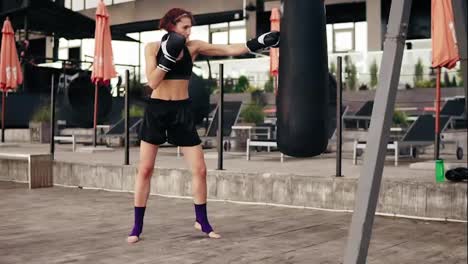 Strong-athletic-female-boxer-in-gloves-exercising-with-a-bag.-She-hits-a-bag-with-her-leg.-Workout-outside.-Female-boxer