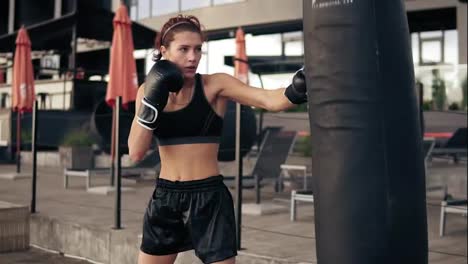 Attractive-athletic-female-boxer-in-gloves-exercising-with-a-bag.-Workout-outside.-Female-boxer-training.-Self-defence-concept
