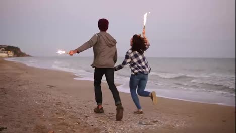 Back-view-of-young-multi-ethnic-couple-holding-burning-sparkling-candles-and-running-by-the-sea-during-sunset.-Slow-Motion-shot