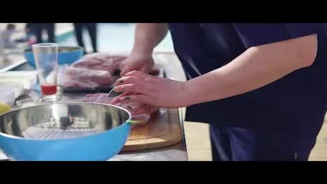 Person's-hands-cutting-a-large-piece-of-raw-meat-using-a-butcher's-knife-on-a-wooden-surface-outside.-Barbeque-preparation.-shot