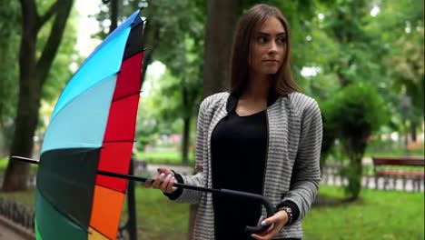 Portrait-of-a-young-attractive-brunette-woman-spinning-her-colorful-umbrella-in-the-city-park-and-looking-in-the-camera