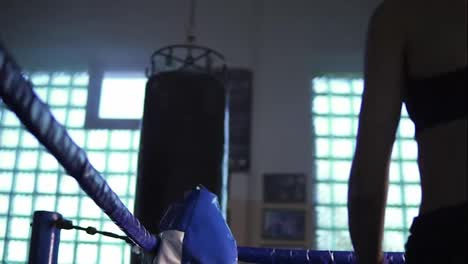 Young-female-boxer-coming-to-the-corner-of-the-boxing-ring-and-resting-with-her-arms-on-the-ring-ropes-in-the-dark-gym