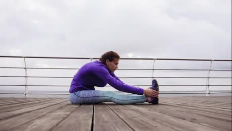 Athletic-woman-stretching-her-legs-sitting-on-the-platform-on-the-beach-by-the-sea-early-in-the-morning.-Training-by-the-beach