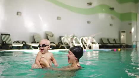 Young-mother-holding-her-little-boy-in-protective-glasses-and-teaching-how-to-swim-in-the-swimming-pool.-Baby-kicking-with-his