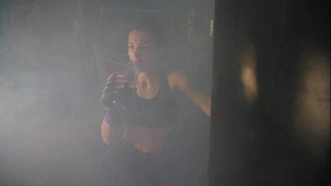 Thirsty-female-boxer-taking-a-break-drinking-from-the-water-bottle-after-training-in-the-dark-gym-with-smoke.-Beautiful-woman