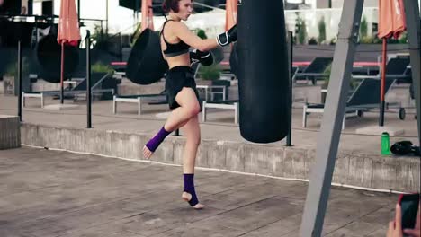 High-pace-video:-Strong-athletic-female-boxer-in-gloves-exercising-with-a-bag.-She-hits-a-bag-with-her-leg.-Workout-outside