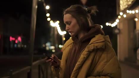 The-girl-in-the-jacket-is-standing-on-the-street-with-lights-in-the-background.-Focused-at-the-phone,-settle-her-hair