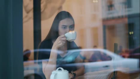 Blick-Von-Der-Straße-Auf-Eine-Junge-Frau,-Die-Im-Café-Tee-Oder-Kaffee-Aus-Der-Weißen-Tasse-Trinkt-Und-Im-Kaffee-Am-Fenster-Sitzt