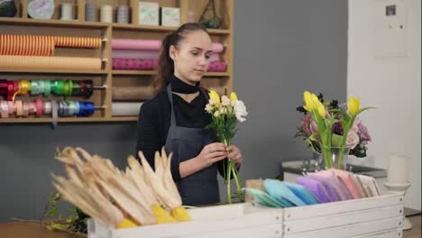 Young-female-florist-arranging-modern-bouquet-at-flowers-shot-standing-by-the-table.-She-combines-creamy-roses-and-yellow-tulips