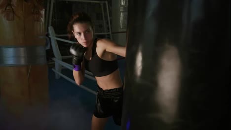 Slow-Motion-shot-of-a-female-boxer-in-gloves-hitting-hard-a-boxing-bag-with-her-fist-while-training-in-a-dark-fitness-studio-with