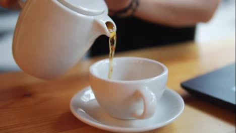Close-up-female-hand-pouring-hot-tea-from-the-teapot-in-the-white-cup.-The-camera-moves-and-shows-a-beautiful-young-girl-with