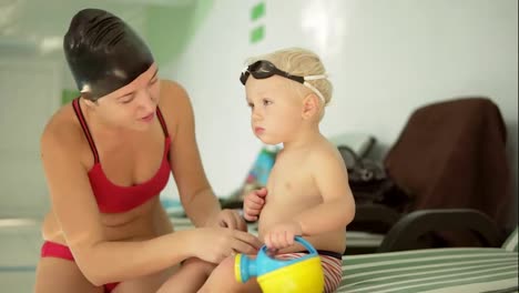 Young-mother-and-her-cute-little-blonde-child-sitting-by-the-swimming-pool-and-preparing-for-the-swimming-lesson.-Happy-mom