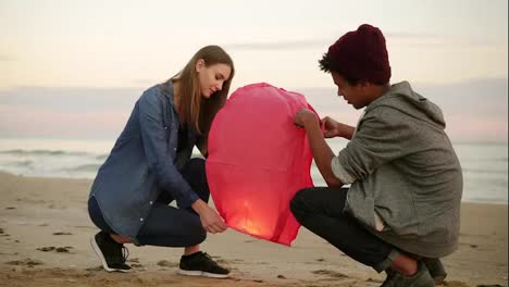 Romantic-date-at-the-beach-during-sunset:-Young-multi-ethnic-couple-holding-red-paper-lantern-before-launching.-Attractive-woman