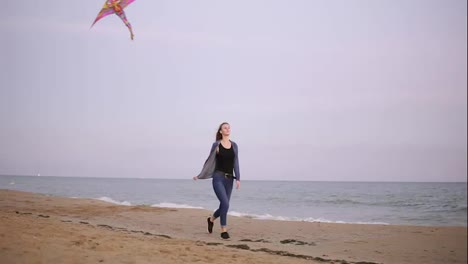 Attractive-young-woman-with-long-hair-holding-colorful-kite-flying-in-the-sky-and-walking-on-the-beach-in-the-evening-during