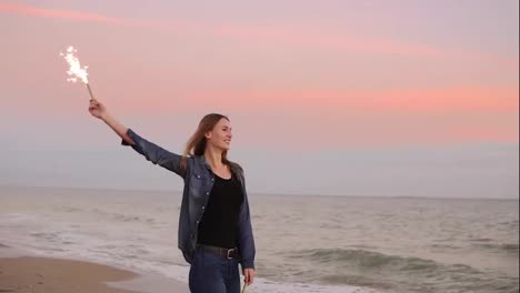 Young-attractive-woman-standing-by-the-sea-during-sunset-and-holding-burning-sparkling-candle.-Slow-Motion-shot