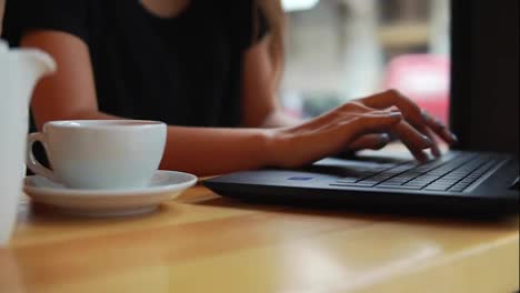 Close-Up-view-of-woman's-hands-typing-on-the-laptop's-keyboard-in-the-coffee-shot-during-a-coffee-break.-White-cup-with-saucer