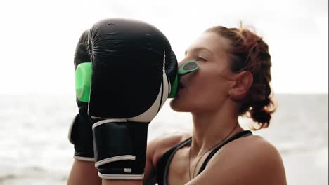 Thirsty-female-boxer-in-gloves-taking-a-break-drinking-from-the-water-bottle-after-training.-Beautiful-woman-training-by-the-sea