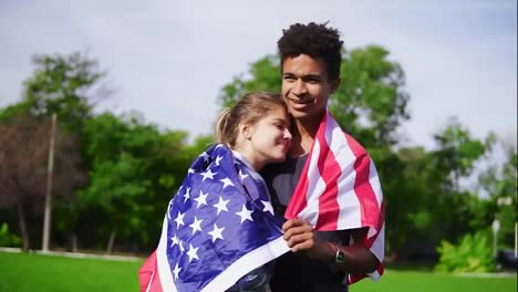 Attractive-multi-ethnic-couple-embracing-each-other-holding-American-flag-on-the-back-standing-in-the-green-field.-Patriotic