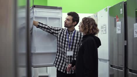 Young-woman-standing-in-front-the-open-door-of-refrigerator-with-male-consultant-discussing-design-and-quality-before-buying-in
