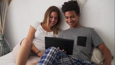 Junges-Multiethnisches-Paar,-Das-Lachend-Filme-Im-Bett-Auf-Einem-Digitalen-Tablet-Anschaut.-Zeitlupenaufnahme