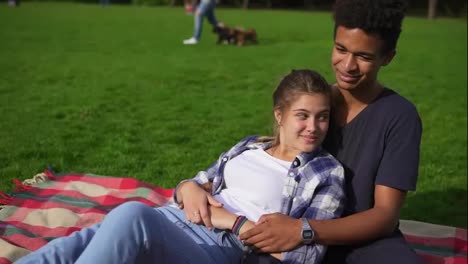 Young-lovely-couple-sitting-on-the-grass-in-park-on-summer-day.-Attractive-man-carefully-embracing-his-girlfriend-and-telling
