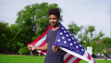 Attraktiver-Afroamerikanischer-Mann-Hält-Die-Amerikanische-Flagge-In-Seinen-Händen-Auf-Dem-Rücken,-Steht-Auf-Der-Grünen-Wiese-Und-Hebt-Sie-Dann-Hoch