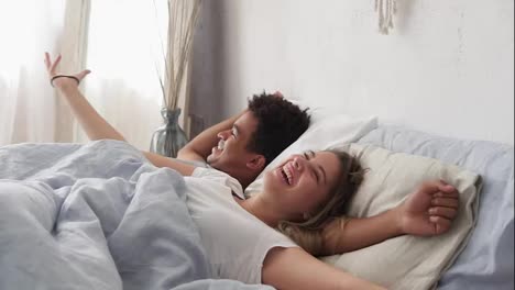 Happy-multiracial-couple-lying-in-bed-and-laughing.-Waking-up-together-in-the-morning.-Attractive-young-man-and-woman-looking-at