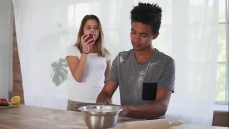 Attractive-african-man-preparing-dough-on-the-wooden-table.-His-girlfriend-is-teasing-him-with-the-apple.-Young-multi-ethnic