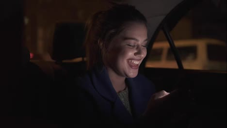 Happy-smiling-girl-riding-in-a-taxi-at-night-sitting-on-the-backseat-and-texting-using-her-smartphone.-Happy-woman-in-taxi