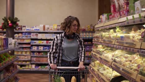 Attractive-girl-customer-with-headphones-on-neck-is-buying-bread-in-bakery-department-is-shot,-looking-on-it-and-putting-in