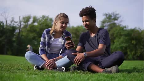 Mixed-race-couple-sitting-on-the-grass-and-selects-pictures-in-their-smartphone.-Young-people-smile-and-laugh.-Cute-friends